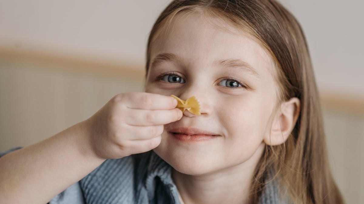 Cute caucasian girl holding bow-tie pasta indoors, smiling happily.